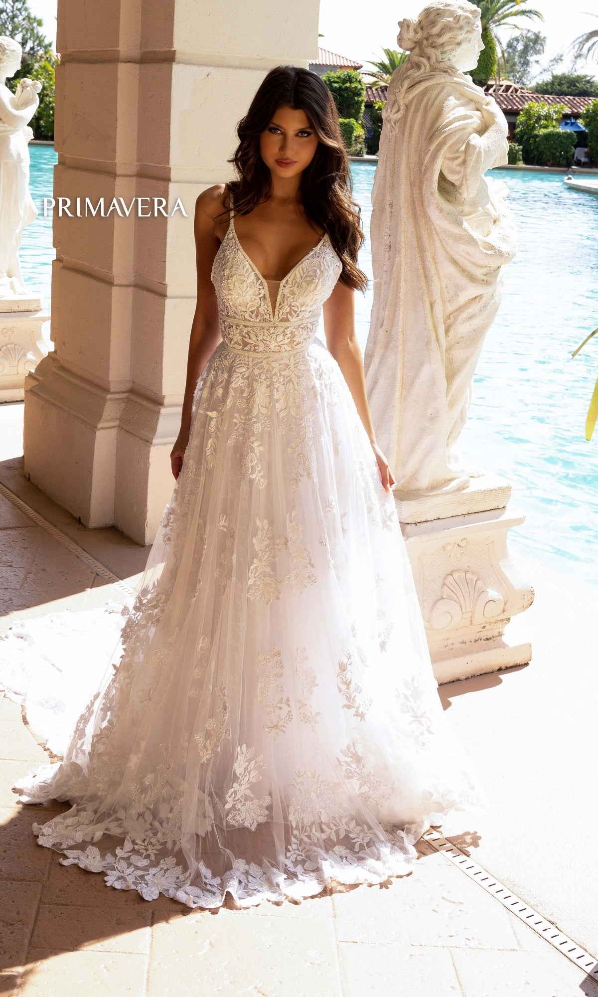 A woman in the Long Wedding Dress 11102 by Primavera—an ivory a-line gown with lace embroidery and a low v-neckline—stands poolside near white pillars and statue, surrounded by water and greenery.